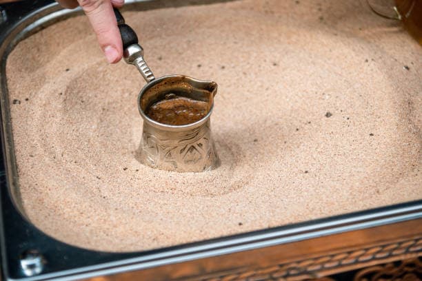 A cezve of Turkish coffee brewing on hot sand in the traditional way